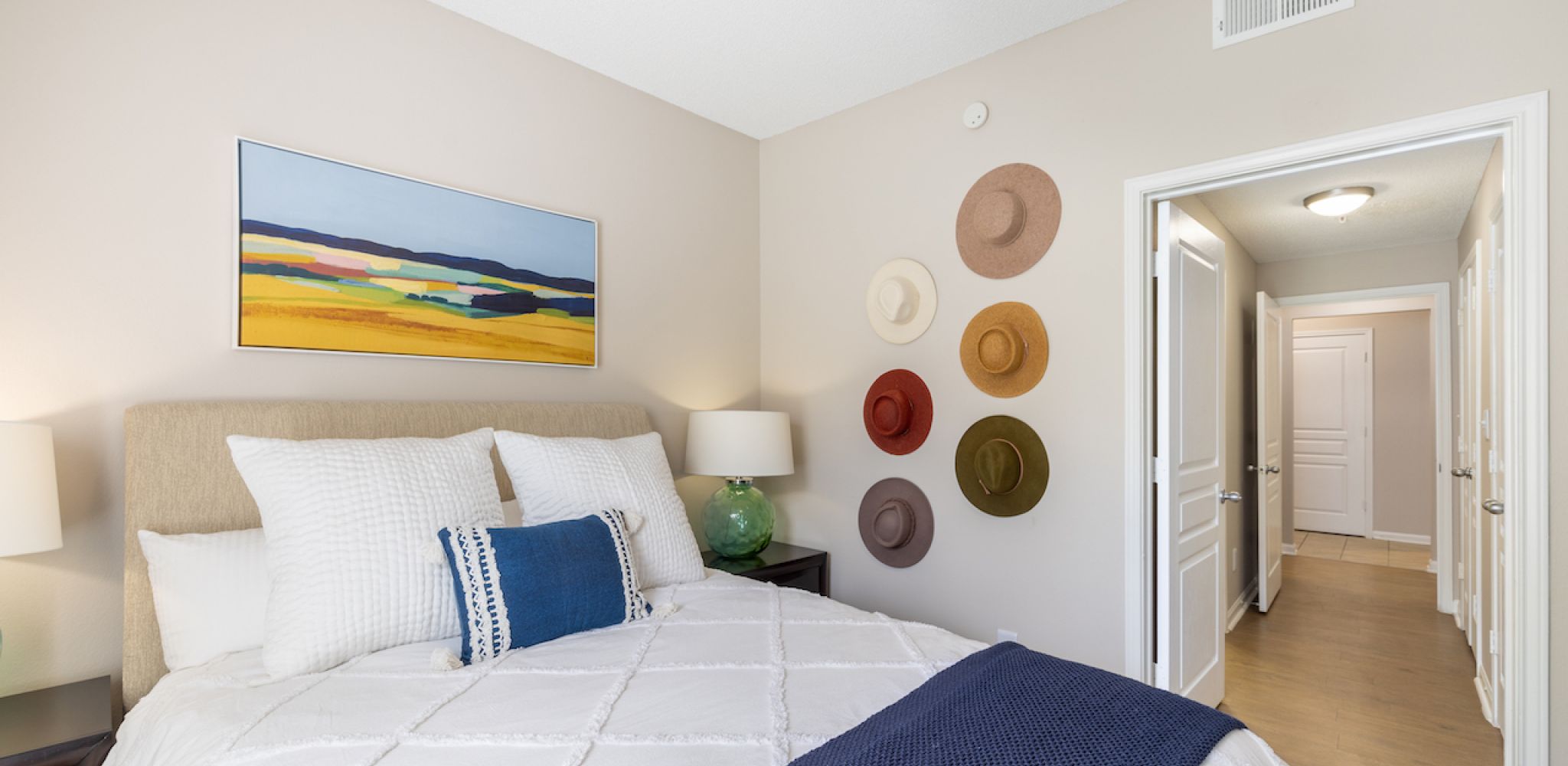 Hawthorne at Lake Heather cozy bedroom featuring a neatly made bed with white and blue accents, stylish decor including a wall display of hats, and a hallway leading to the rest of the apartment.